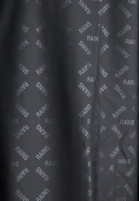 Un tissu gris foncé présente un motif diagonal répétitif du mot "RAINS" en gris clair, mettant en valeur une texture lisse et une finition mate.