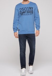 Blauer Pullover mit schwarzem Schriftzug auf der Vorderseite, kombiniert mit dunkler Jeans und weißen Turnschuhen. Durchgehende gerippte Bündchen an Ärmeln und Saum.