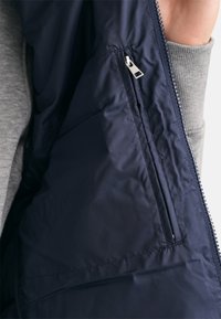 Interior de chaqueta aislante azul marino con una textura suave, bolsillo lateral con cremallera y forro de punto gris en el cuello y los puños.