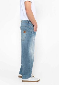 Jean en denim bleu clair avec une coupe ample, présentant des coutures distinctives, une étiquette en cuir sur la poche arrière, et des poches latérales.