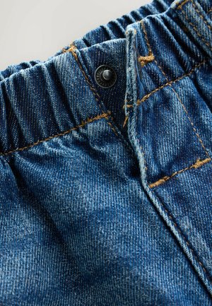 Tejido de denim azul con una cintura fruncida y un botón de metal negro. La costura visible en marrón crea textura y detalle.