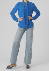 Niebieska bluzka z długim rękawem z falbaniastym przodem, w połączeniu z jasnoniebieskimi spodniami typu wide-leg i niebieskimi sandałami z odkrytymi palcami, ozdobionymi kwiatowym akcentem.