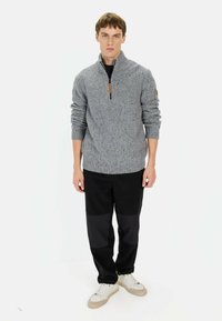 Grijze gebreide pullover met hoge kraag en ritssluiting, voorzien van geribbelde manchetten en een textuurpatroon. Draagt een zwarte broek en sneakers.