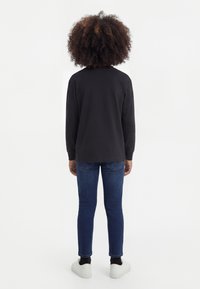 Kiharatukkainen henkilö seisoo selin, yllään musta pitkähihaisen paita, siniset farkut, mustat sukat ja valkoiset lenkkarit.