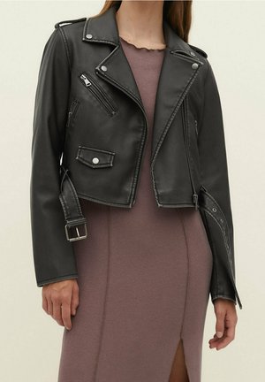 Femme portant une veste motard courte en cuir noir avec des fermetures éclair et des boucles argentées, par-dessus une robe moulante mauve avec une fente devant.