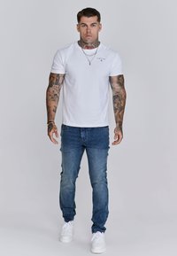 T-shirt bianco a maniche corte con logo piccolo, abbinato a jeans di denim blu medio e sneaker bianche. Presenta tatuaggi sulle braccia.