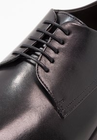 Chaussure à lacets en cuir noir, présentant une texture lisse, un bout arrondi et six œillets. Finition brillante avec des détails de couture subtils.