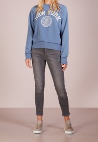 Felpa blu con testo e logo "NEW YORK", abbinata a jeans slim-fit grigi e sneakers beige. Tessuto morbido con polsini a coste.