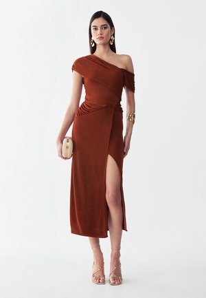 Frau trägt ein rostfarbenes Ein-Schulter-Kleid mit seitlichem Schlitz, hält eine goldene Clutch und trägt goldene Ohrringe, Armband sowie Riemchenabsätze.