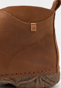 Hnědá kožená kotníková botka s texturovaným povrchem, šitým detailem kolem vrchu a malou koženou nášivkou na zadní straně.