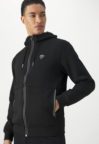 Felpa nera con zip realizzata in tessuto morbido, con cappuccio, tasche frontali con cerniere e una toppa con logo sul petto.