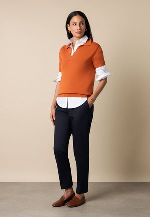 Frau steht und trägt einen orangefarbenen Kurzarm-Pullover über einem weißen Hemd, schwarze Hose und braune Loafers, schaut nach links.