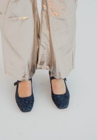 Ballerine glitter blu navy con punta quadrata e cinturino alla caviglia, abbinate a pantaloni larghi metallici lucidi che riflettono la luce.