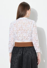 Weißes Spitzen-Crop-Top mit langen Ärmeln, das florale Muster zeigt. Kombiniert mit einem schwarzen Rock und einem braunen Taillenbündchen im Mittelteil.
