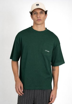 Mężczyzna w beżowej czapce, ciemnozielonej koszuli z krótkim rękawem i przednią kieszenią oraz ciemnych spodniach w prążki, stoi na białym tle.