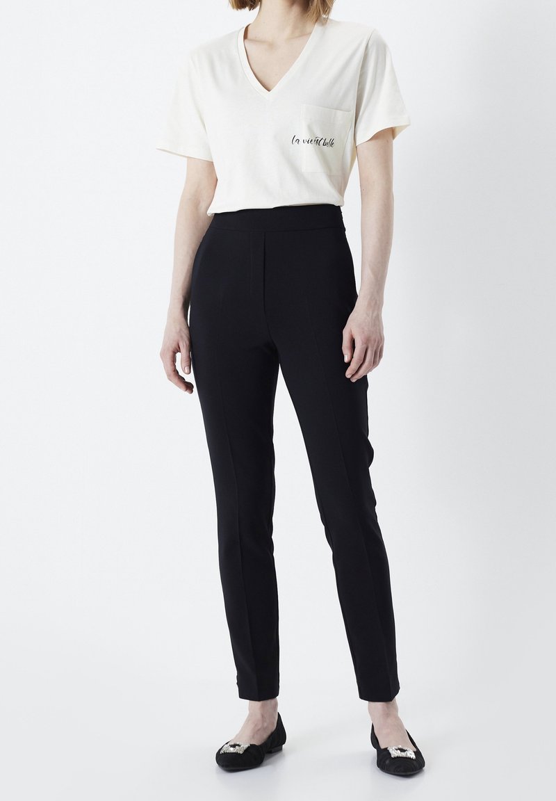 Femme portant un t-shirt blanc à col en V avec une poche et un texte, associé à un pantalon noir slim et des ballerines noires ornées d'embellissements argentés.