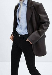 Blazer marrón de gran tamaño con hombros estructurados, superpuesto a una camisa de botón azul claro, combinado con jeans slim-fit negros. Texturas suaves.