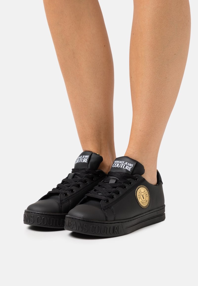 Jambes portant des baskets noires Versace Jeans Couture avec des lacets noirs et un écusson logo doré, vues sur un fond clair uni.