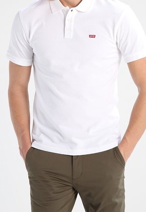 Mężczyzna w białej koszulce polo Levi's z trzema guzikami i oliwkowozielonych spodniach, z rękami w kieszeniach, na jednolitym jasnym tle.