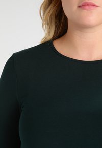 Ciemnozielona bluzka z długim rękawem i okrągłym dekoltem, wykonana z gładkiego materiału, charakteryzująca się dopasowanym krojem i minimalistycznym designem.