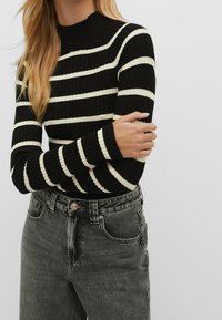 Haut à manches longues noir à côtes avec des rayures crème ; design col montant. Porté avec un jean en denim gris taille haute avec fermeture à bouton.