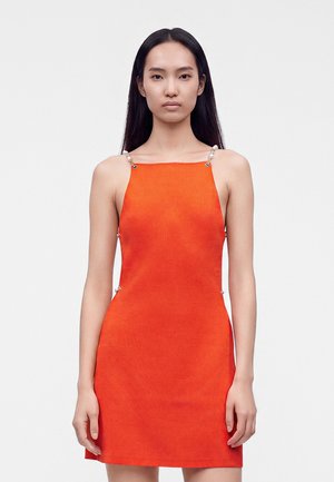 Jeune femme aux longs cheveux foncés portant une robe orange sans manches avec des bretelles fines ornées de perles, debout devant un fond blanc uni.
