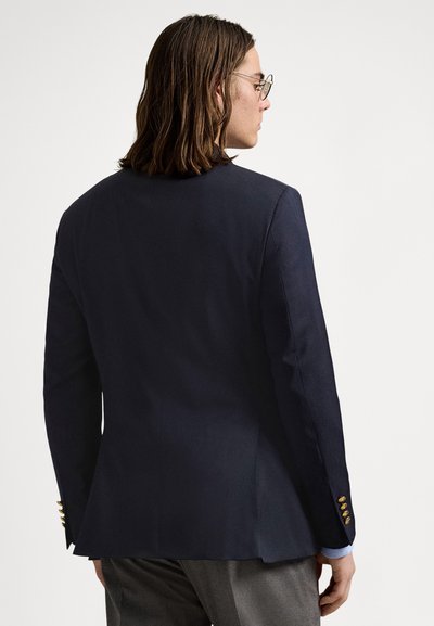 Blazer azul-marinho com corte ajustado, apresentando ombros estruturados, detalhes de botões dourados nos punhos e textura de tecido suave.