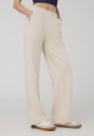 Pantaloni della tuta beige chiaro con un elastico in vita, tasche laterali e una vestibilità rilassata, abbinati a sneakers bianche e grigie su uno sfondo neutro.