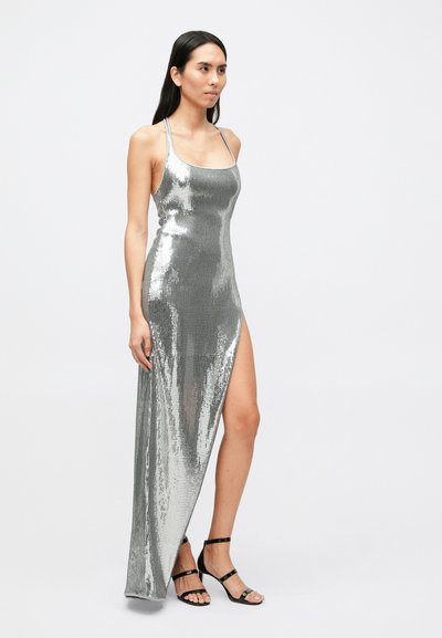Strieborné flitrové šaty s vysokým rozparkom, tenkými ramienkami a priliehavým strihom. Hladká textúra a lesklý povrch zvýrazňujú vizuálny dojem.