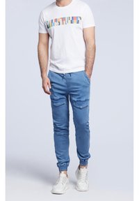 Camiseta blanca de manga corta con un logo horizontal multicolor; pantalones jogger azul claro con puños elásticos y bolsillos frontales; zapatillas blancas.