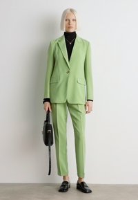 Hellgrüner maßgeschneiderter Anzug mit einem einzigen Knopf. Kombiniert mit einem schwarzen Rollkragenpullover und schwarzen Slippern. Hält eine schwarze Handtasche und steht an einer weißen Wand.