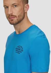 Blauwe katoenen t-shirt met korte mouwen en een ronde hals. Heeft zwarte bedrukte tekst aan de linkerkant. Gladde textuur, standaard pasvorm.