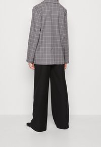 Harmaa ruudullinen bleiseri suoralinjaisella leikkauksella, jossa on näkyvä sauman yksityiskohta takana. Yhdistetty leveiden mustien housujen ja mustien lenkkareiden kanssa.