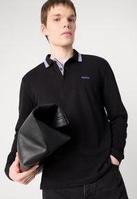 Camisa polo preta de manga longa com colarinho em contraste, apresentando detalhes prateados e roxos, acompanhada de uma carteira de mão preta de couro.