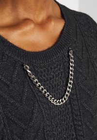 Pull noir en maille avec motif à torsades texturé, orné d'un accessoire chaîne métallique argentée au niveau de l'encolure.