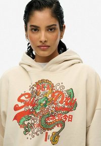 Hoodie beige présentant un design de dragon brodé coloré avec des accents rouges et verts. Coupe décontractée avec une capuche à cordon.