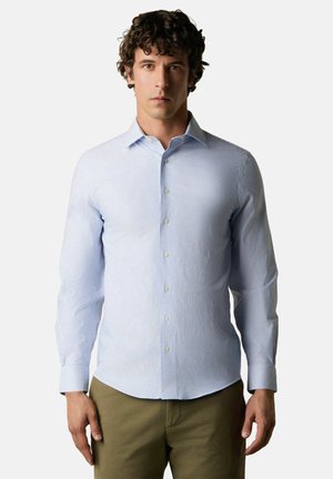 Man met donker krullend haar, gekleed in een lichtblauw, dun gestreept overhemd met knopen en olijfgroene broek, staand tegen een effen achtergrond.