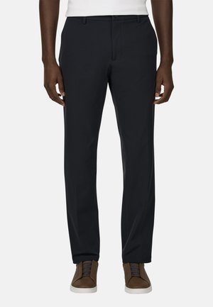 Mann trägt schwarze Slim-Fit-Hose mit Seitentaschen und braune Sneakers mit weißen Sohlen, steht vor einem einfarbigen Hintergrund.