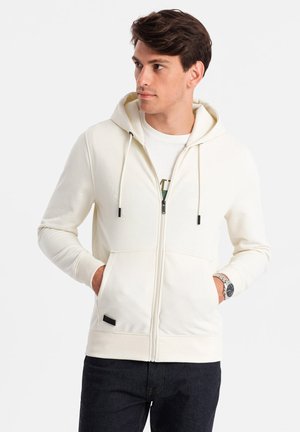 Cream zip-up hoodie with a hood, front pockets, and drawstrings. Textured fabric with a black label at the hem and ribbed cuffs.