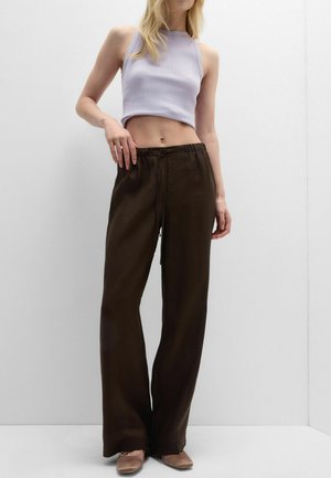 Femme portant un haut court sans manches violet clair, un pantalon large marron foncé et des chaussures plates marron sur un fond blanc uni.