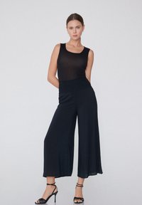 Haut sans manches noir associé à un pantalon large noir. La tenue présente une taille ajustée et se déploie de manière fluide, complétée par des talons noirs à lanières.