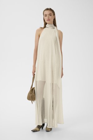 Kvinde iført ærmeløs cremefarvet maxi-kjole med høj hals og små prikker, holder en beige taske og har slangeskinds-ankelstøvler på.