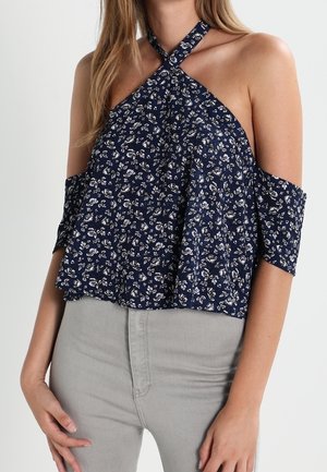 Femme portant un haut dos nu bleu marine avec imprimé floral blanc et manches épaules dénudées, associé à un pantalon gris clair.