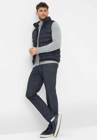 Marineblauw nylon puffer vest over een grijze gebreide trui, gecombineerd met donkere, gestructureerde broeken en marineblauwe sneakers met een witte zool.