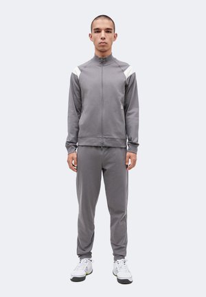 Ung mand står vendt fremad iført en grå træningsdragt med hvid skulderdetalje og hvide sneakers på en ensfarvet baggrund.