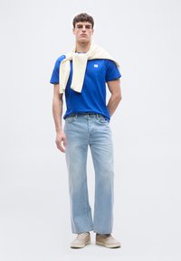 Νεαρός άνδρας που φορά μπλε μπλούζα, ανοιχτόχρωμο τζιν, μπεζ αθλητικά παπούτσια, με κρεμ πουλόβερ περασμένο και δεμένο γύρω από τους ώμους του.