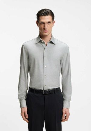 Mand med tilbageglattet hår iført en lysegrå skjorte med lange ærmer, knappet op og stoppet ned i sorte bukser med et sort bælte.