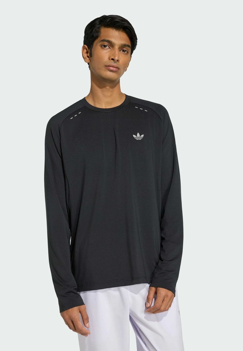 Jeune homme portant un t-shirt noir à manches longues Adidas avec logo sur la poitrine, debout devant un fond clair uni.