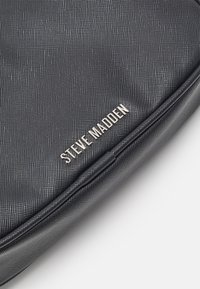Black textured handbag with silver "Steve Madden" logo, featuring a smooth finish and rounded edges. Compact and minimalist design.