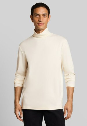 Homme portant un pull à col roulé crème avec les manches retroussées et un pantalon noir, debout devant un fond gris uni.